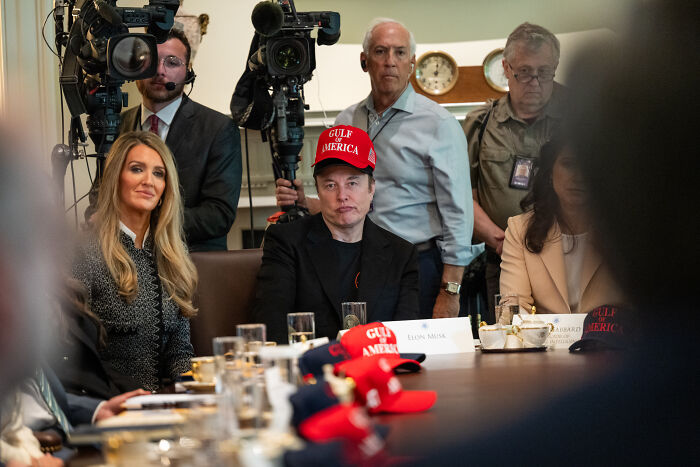 Elon Musk at a meeting wearing a Gulf America hat, surrounded by people and media crew, discussing government waste.