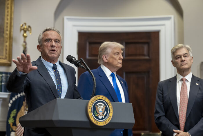 Three men at a podium with the presidential seal, featuring Trump discussing a well-known overweight neurotic pal's fat shot abroad.