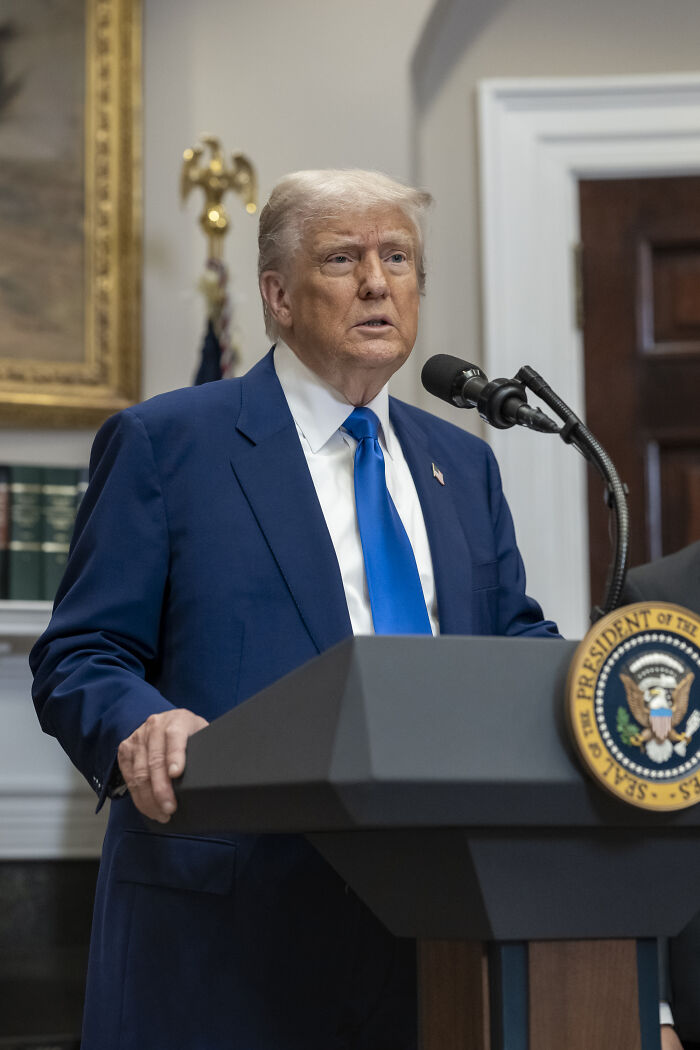Donald Trump speaking at a podium during a public address about his overweight and neurotic pal’s fat shot abroad.
