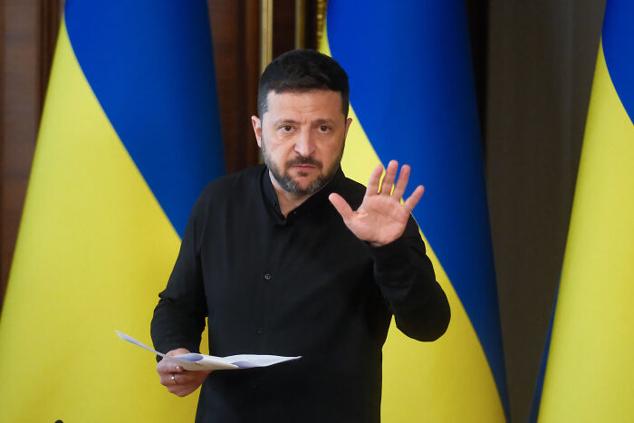 Volodymyr Zelenskyy speaking with raised hand, Ukrainian flags in background amid Russian attack on Ukraine situation.