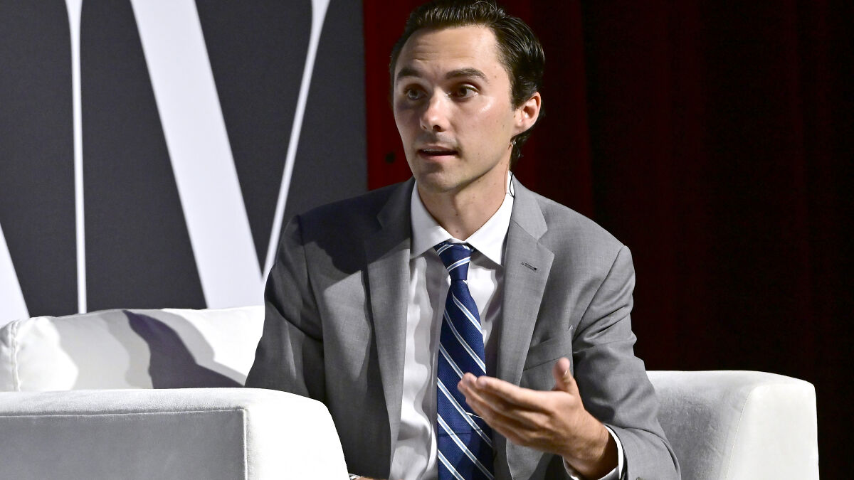 David Hogg speaking at an event, wearing a gray suit and blue striped tie, representing DNC committee news.