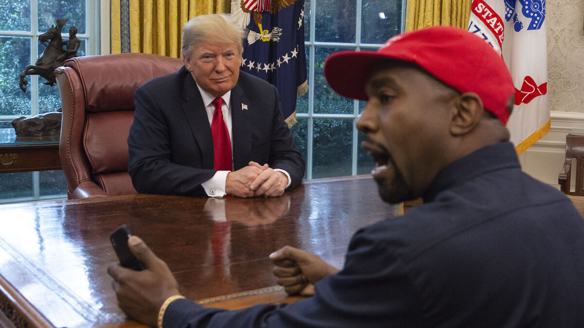 Donald Trump sitting at the Resolute Desk listening while Kanye West speaks during a meeting in the Oval Office