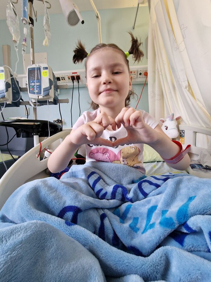Seven-year-old girl in hospital bed making a heart shape with hands while receiving cancer treatment in Iran.