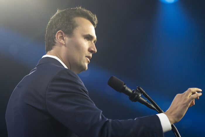 Conservative pundit in a suit speaking at a microphone, with blue background, amid critics mocking his US town video.