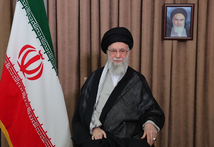 Iran’s Supreme Leader seated near the national flag, delivering a serious address amid rising tensions with the U.S. and Israel.