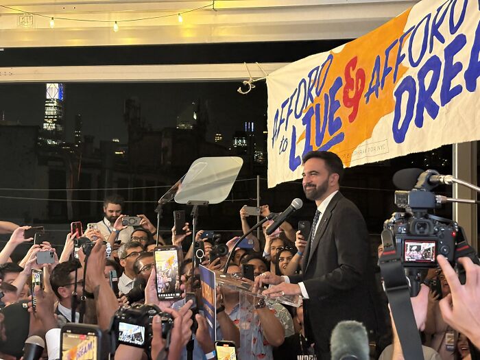 Democratic Socialist Zohran Mamdani speaking at NYC mayoral primary victory event with supporters capturing the moment on phones.