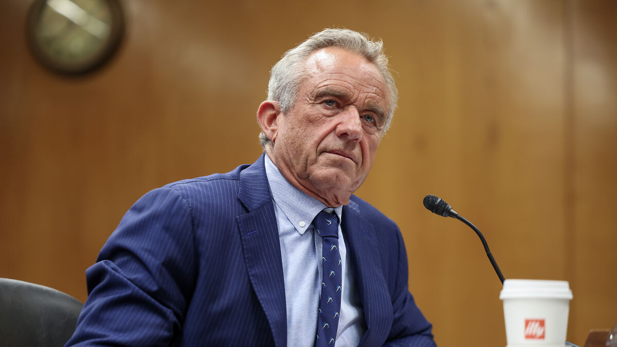 Robert F. Kennedy Jr. speaking at a hearing, wearing a blue suit with a microphone and coffee cup nearby.