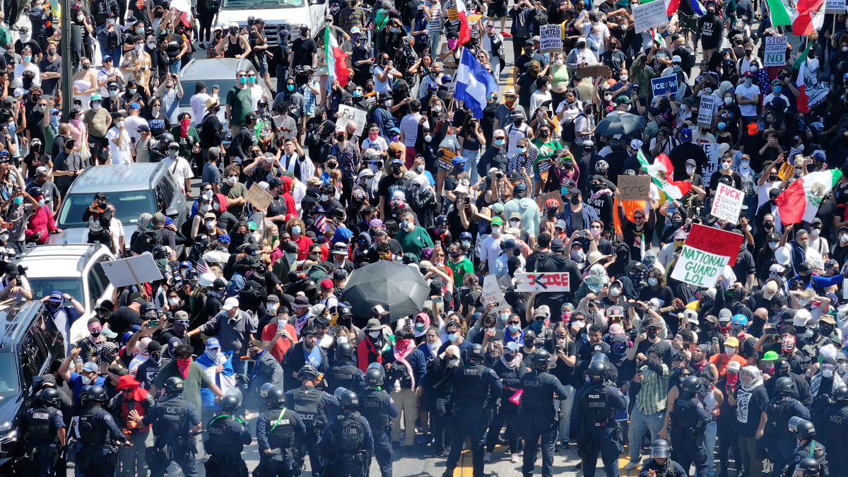 Large crowd protesting with signs and police officers in riot gear during National Guard control dispute in California.