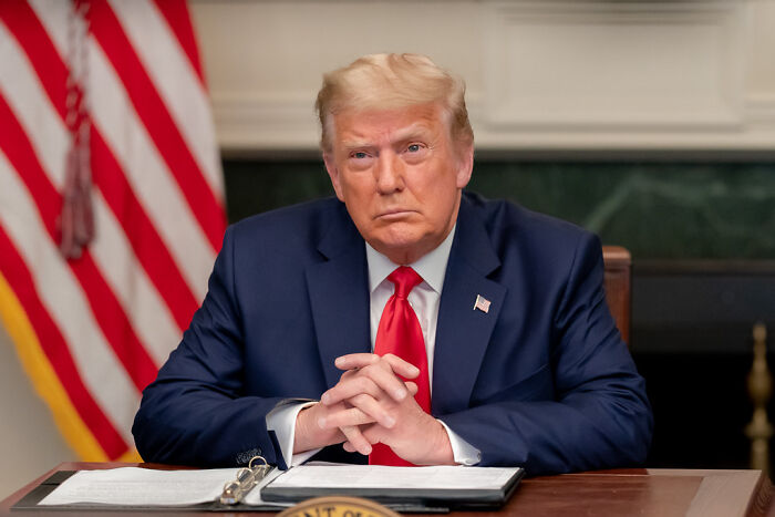 Donald Trump seated at a desk with folded hands, related to Trump Penned Epstein Birthday Note legal action news.