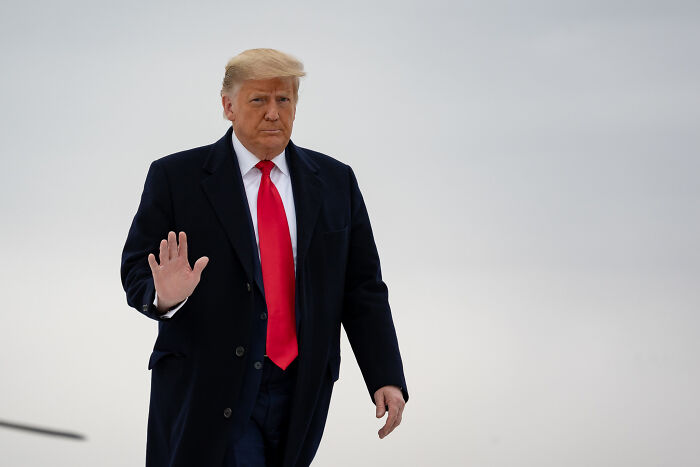 Former President Donald Trump in a dark coat and red tie, walking outdoors with a neutral gray sky background. Former President Donald Trump in a dark coat and red tie, walking outdoors with a neutral gray sky background.