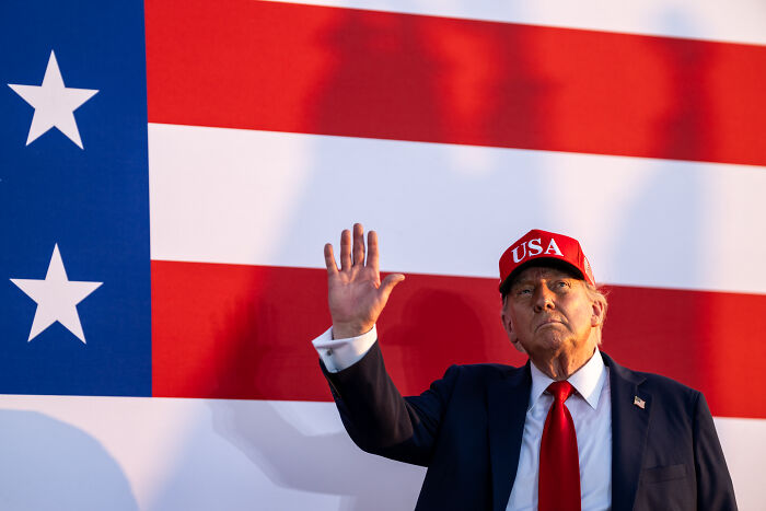 Former President Trump wearing a red USA hat, raising his hand in front of a large American flag backdrop.