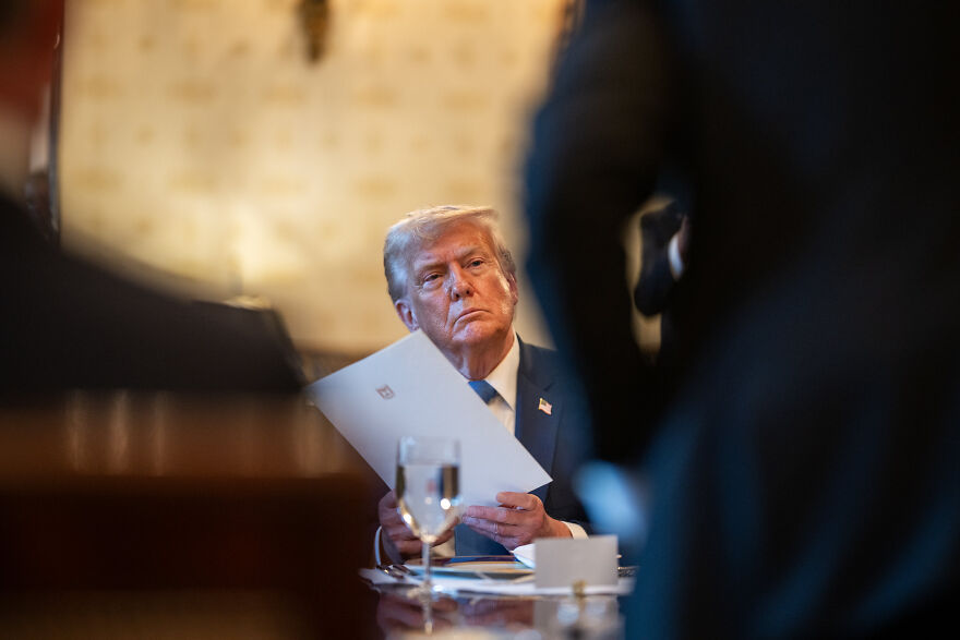 Donald Trump seated at a table holding papers, with blurred figures in foreground, relating to Epstein files and Republicans blocking release.