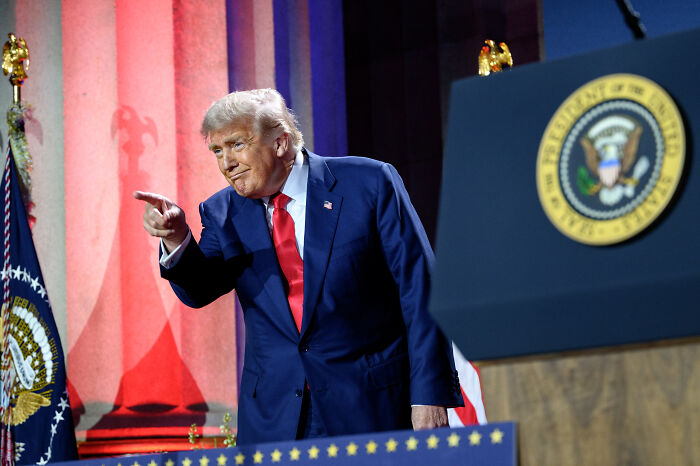 Donald Trump in a navy suit and red tie pointing during a speech at an event with presidential seals nearby.