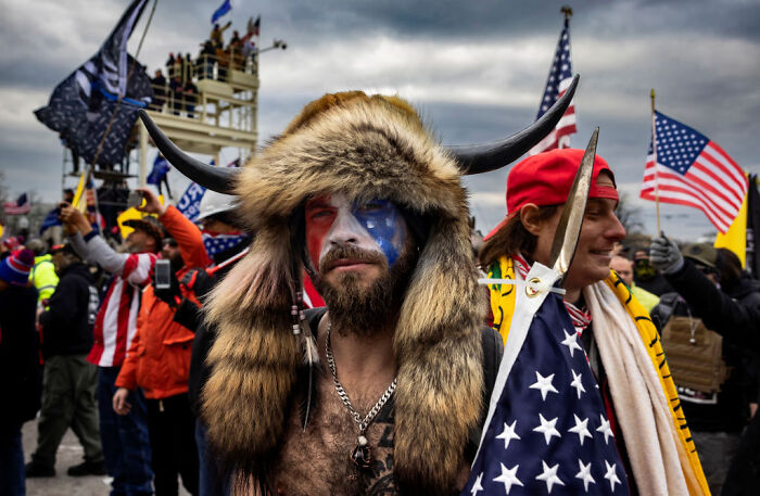 Man dressed as QAnon Shaman wearing a horned fur hat and face paint among crowd holding American flags.