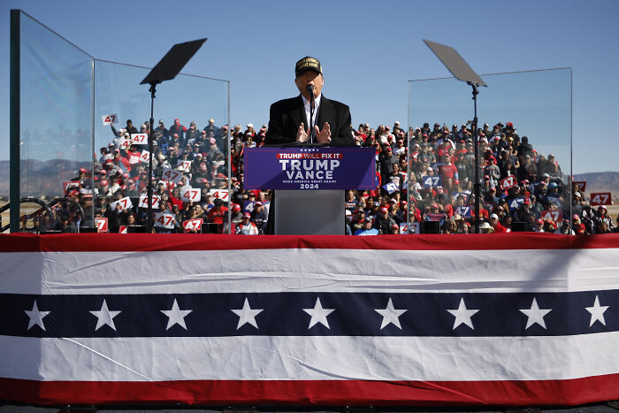 Donald Trump speaking at a rally with crowd holding signs, emphasizing Beyoncé prosecution over Kamala Harris support.