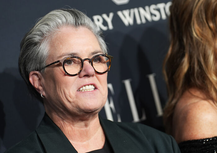 Ellen wearing glasses and a dark blazer, speaking at an event, stunning fans by siding with Rosie O’Donnell.