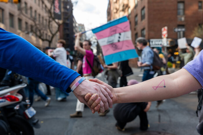 Two people holding hands in a city protest with a transgender pride flag, representing the impact of Trump’s anti-trans agenda.
