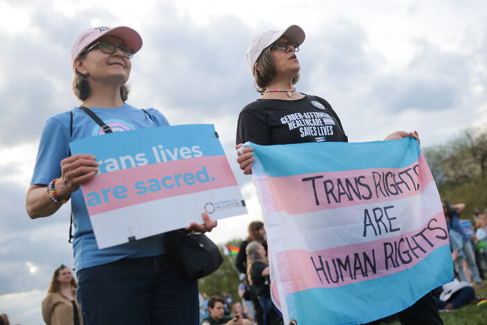 Two protesters holding signs advocating for trans rights and highlighting the impact of Trump’s anti-trans agenda.