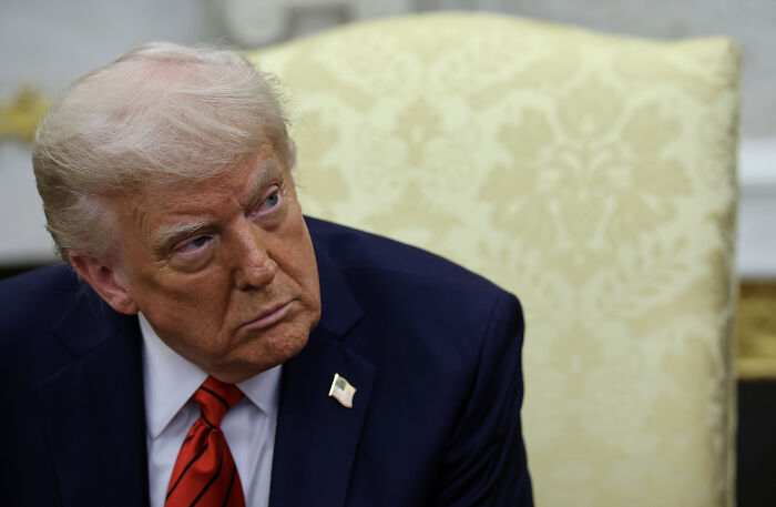 A man in a dark suit with a red tie sitting in an ornate room, reflecting on new tariffs affecting European cars.