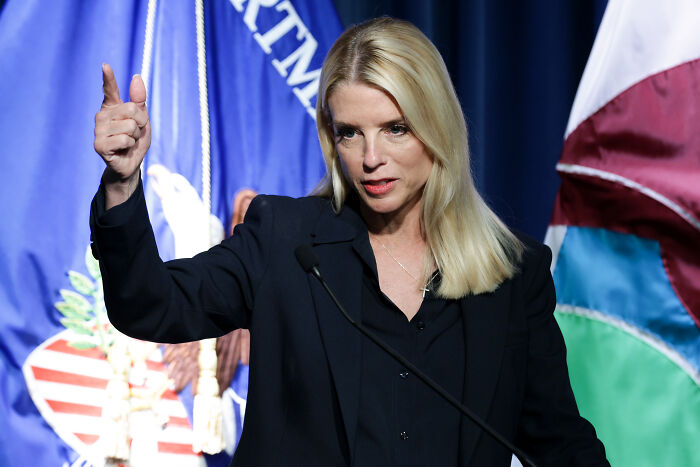Woman with blonde hair speaking at podium with DOJ and US flags behind, addressing immigration sanctuary laws.