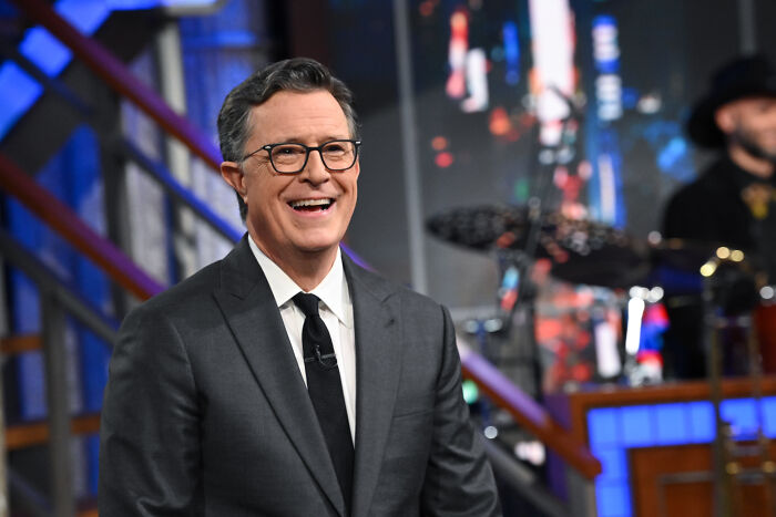 Stephen Colbert smiling on a TV set, dressed in a suit and glasses, related to Trump’s quest to sue on TV.