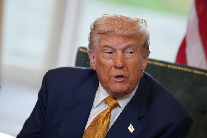 A man in a dark suit and gold tie, seated indoors, discussing U.S. and EU trade deal tariffs on European goods.