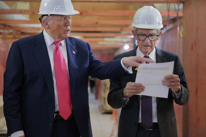 Federal Reserve Chair Jerome Powell wearing a hard hat reviewing documents during a construction site visit with Donald Trump.