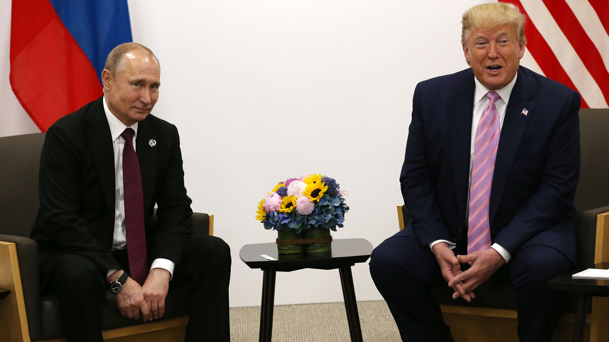 Vladimir Putin and Donald Trump seated in a formal meeting room with flags and a flower arrangement on a table.