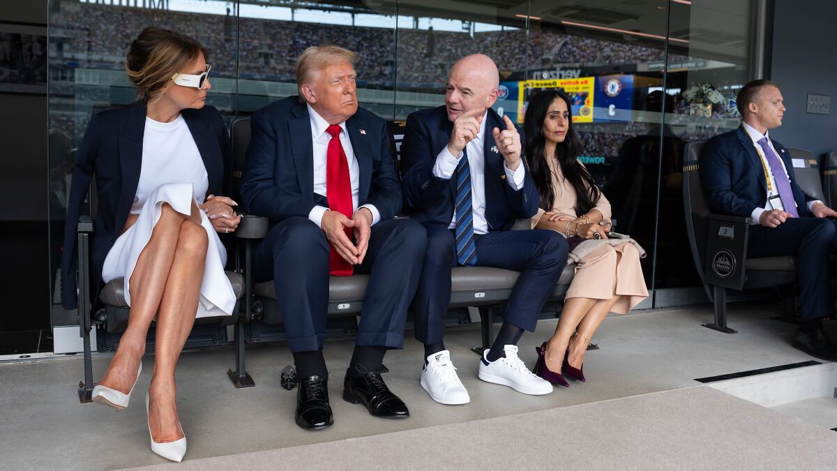 Donald Trump sitting with others at an event, showing visible swollen legs and wearing a dark suit with a red tie.