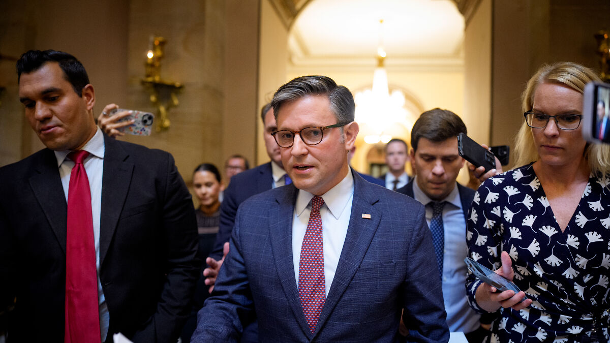 Speaker Mike Johnson in a suit and glasses walking with aides and reporters amid growing bipartisan pressure on Epstein files.