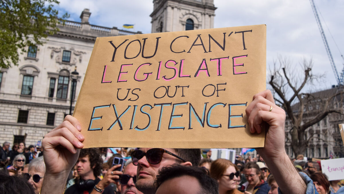 Protesters holding a sign against anti-trans legislation impacting lives during a crowd demonstration outdoors.