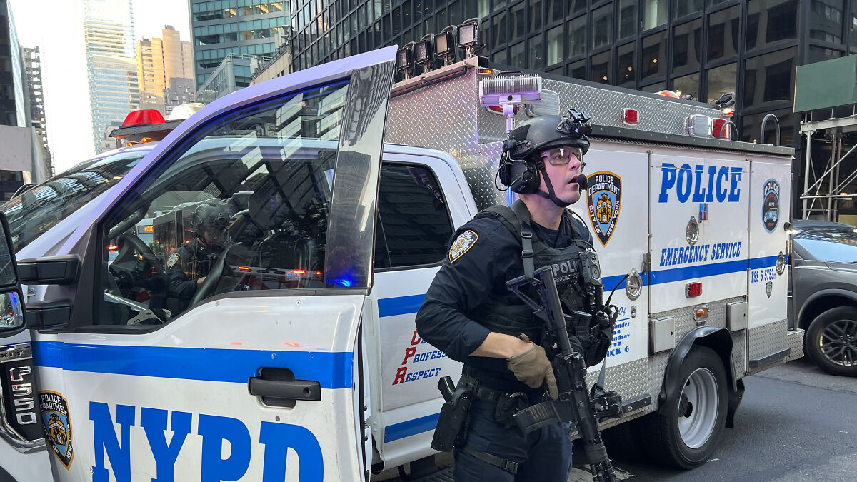 NYPD officer in tactical gear standing beside emergency service vehicle during New York shooter investigation.