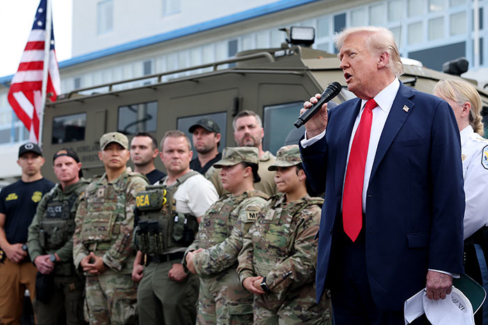 Donald Trump speaking at event surrounded by military personnel and law enforcement, showing a strong leadership stance.