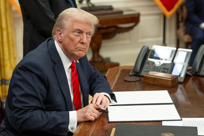 Donald Trump sitting at a desk in the Oval Office, focused and holding a pen, related to Trump praising Sydney Sweeney.