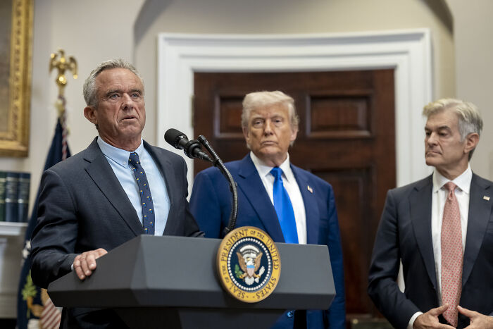 Man in suit speaking at presidential podium with two men standing behind, related to mRNA vaccine development funding.