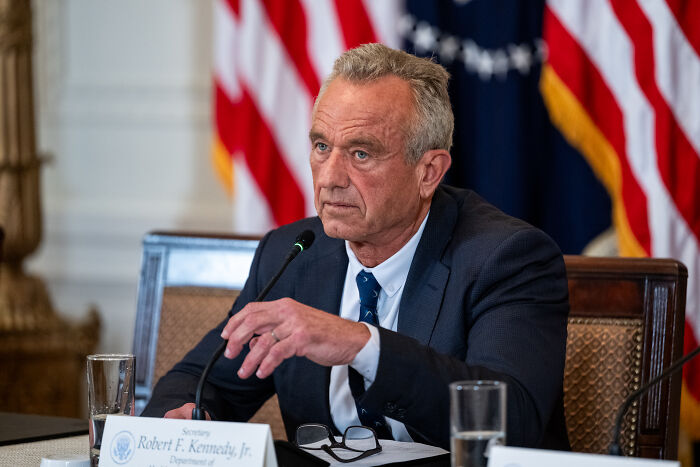 Robert F Kennedy Jr. at a formal meeting, speaking into a microphone with American flags in the background.