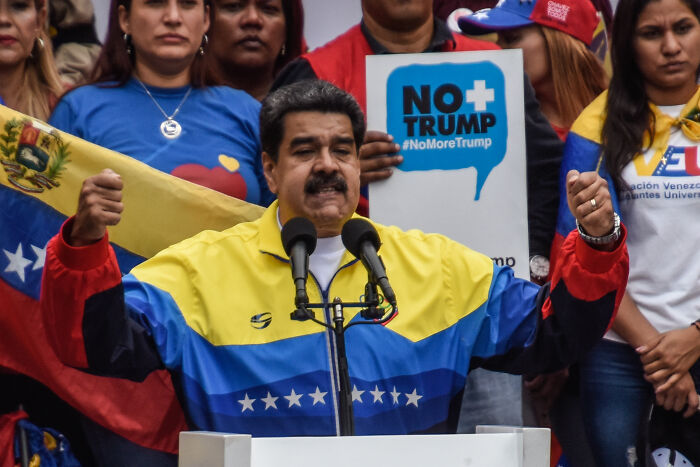 Venezuelan President Nicolás Maduro speaking at a rally with supporters holding flags and anti-Trump signs.