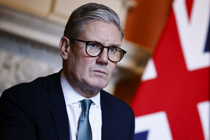 Man in glasses and suit speaking in front of a British flag about major countries recognizing Palestine and future implications