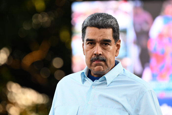 Nicolás Maduro wearing a light blue shirt, looking serious outdoors with blurred background and bokeh lights.