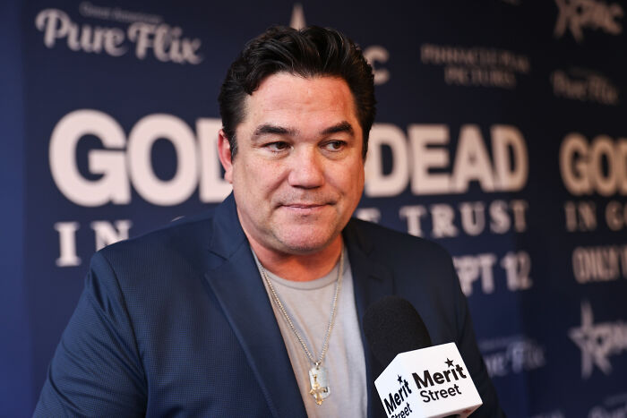John Leguizamo speaking at an event, wearing a dark blazer and necklace, addressing Dean Cain and ICE controversy.