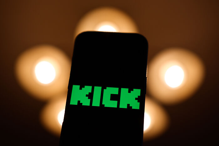 Smartphone screen showing Kick app logo with blurred lights in background highlighting latest streamer tragedy keywords.