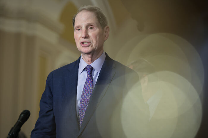 A senator speaking at a podium, addressing the Epstein files controversy in a formal indoor setting.