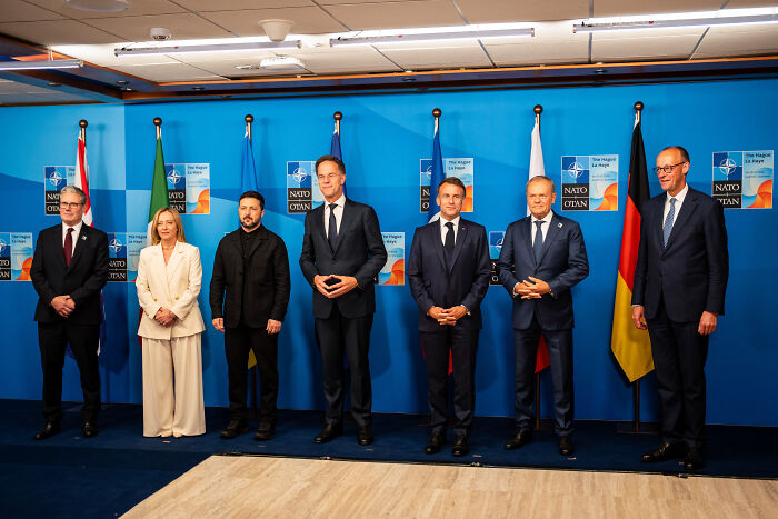 Group of political leaders standing in front of NATO and national flags discussing Ukraine war and Zelenskyy’s role.