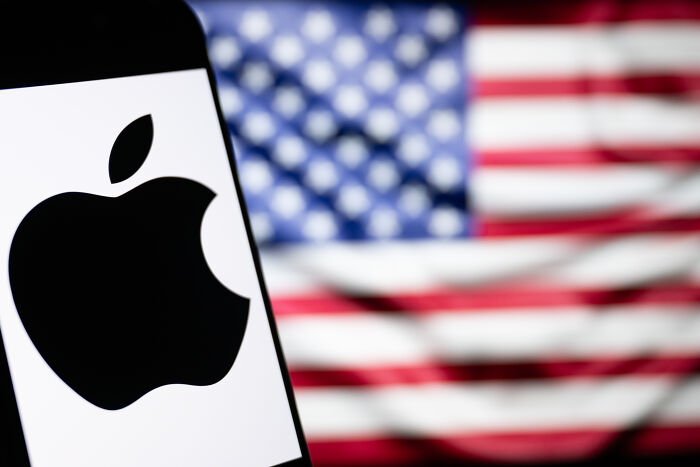 Apple logo displayed on a smartphone screen with a blurred American flag in the background representing Apple’s Tim Cook and White House.