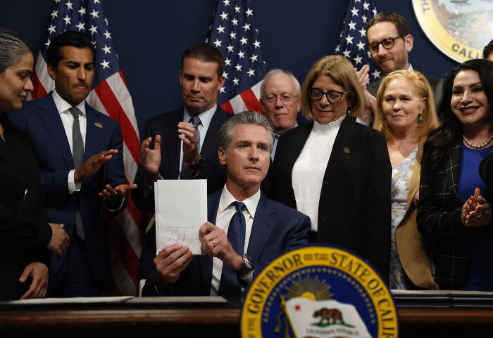 California Governor signing the redistricting plan, surrounded by officials and U.S. flags in a formal setting. California Governor signing the redistricting plan, surrounded by officials and U.S. flags in a formal setting.