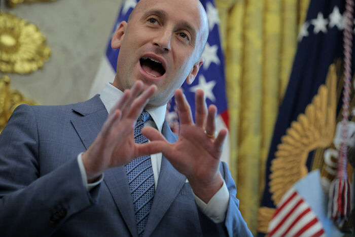 Stephen Miller speaking passionately in a suit with American flags in the background, highlighting Gavin Newsom troll game.