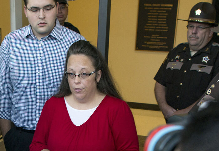 Woman in red speaking to media, accompanied by men and law enforcement, amid a same-sex marriage legal battle.