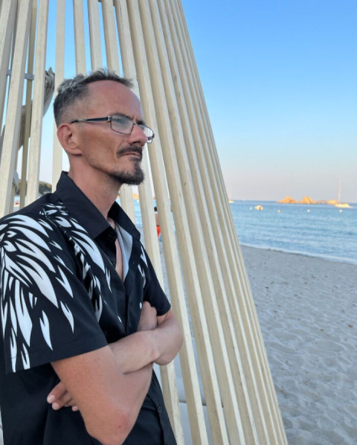 Man with glasses in a black patterned shirt standing on the beach, reflecting on latest streamer tragedy and toxic online challenges.