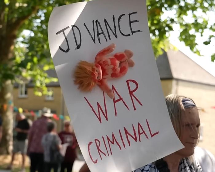 Protester holds sign calling JD Vance a war criminal amid villagers’ fury over disabled parking takeover.