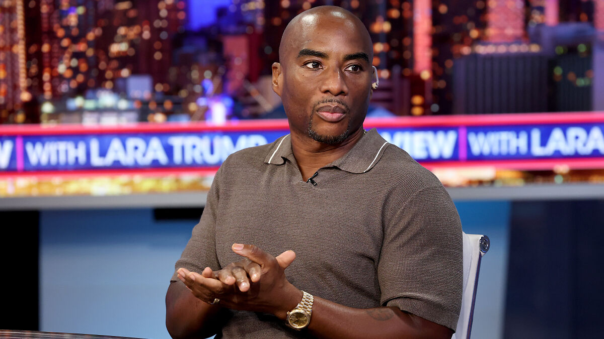 Charlamagne Tha God speaking on a TV show set with a cityscape backdrop in a casual brown shirt and gold watch.